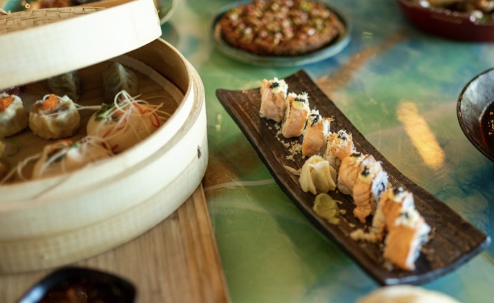 A beautifully arranged spread featuring sushi rolls, dim sum in a bamboo steamer, and vibrant Asian-inspired dishes on a colorful tabletop.