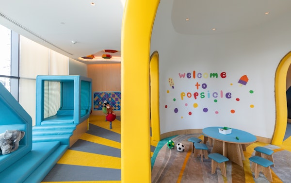 A colorful children's play area featuring a slide, a play wall, and a welcoming "Welcome to Popsicle" sign on the wall, with vibrant yellow and blue accents.