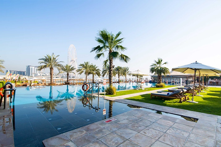 A serene view of a poolside area with palm trees, sunbeds, and an umbrella, with a Ferris wheel in the background.