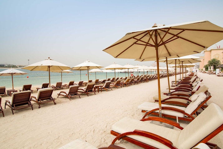 Sofitel The Palm A beach with rows of wooden lounge chairs and umbrellas facing the water.