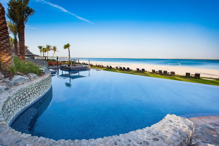 JA The Resort A stunning infinity pool overlooking a sandy beach and the ocean, surrounded by palm trees and lounge chairs under a clear blue sky