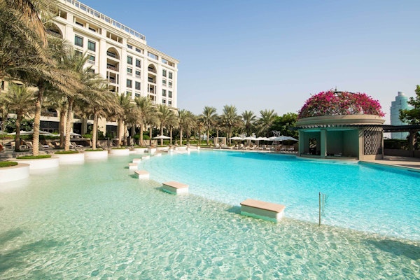 A serene pool area surrounded by palm trees and a luxury hotel, with clear blue water and a floral-decorated pavilion