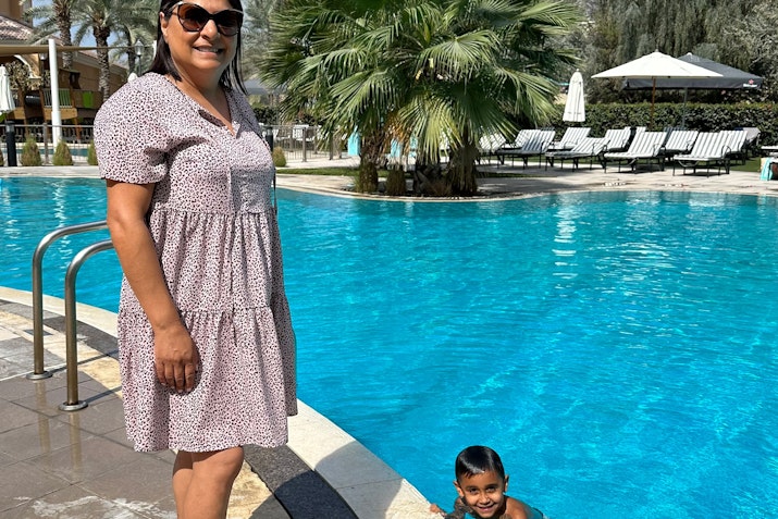 A woman standing by the pool, with a child swimming nearby, surrounded by palm trees and lounge chairs.