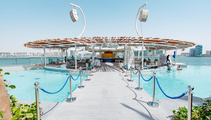 Café del Mar, Abu Dhabi Luxurious pool deck leading to a modern bar area with a wooden canopy, surrounded by water and lounging spaces.