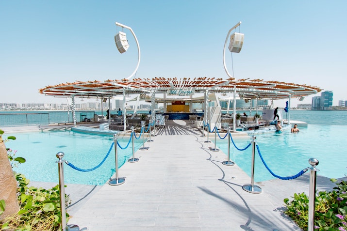 Luxurious pool deck leading to a modern bar area with a wooden canopy, surrounded by water and lounging spaces.