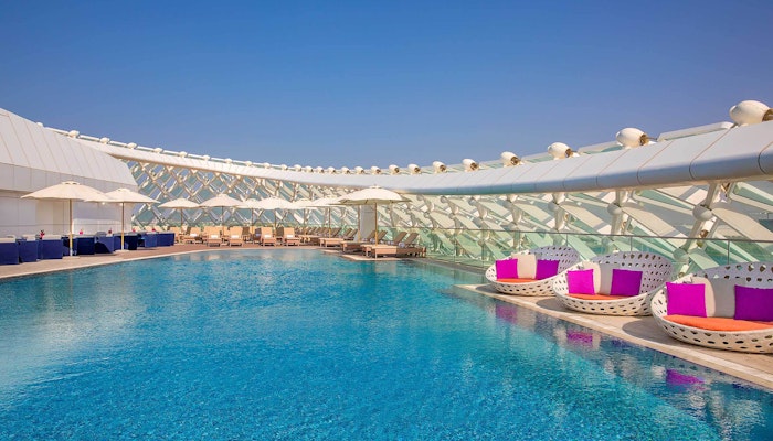 W Abu Dhabi Yas Island – WET Deck A rooftop pool with lounge chairs and circular seats with purple cushions, surrounded by a modern structure and clear blue sky.