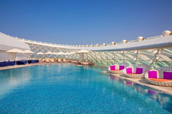 A rooftop pool with lounge chairs and circular seats with purple cushions, surrounded by a modern structure and clear blue sky.