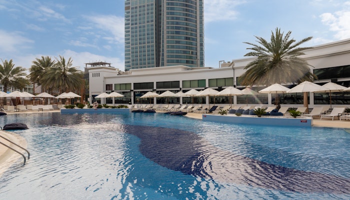 West Bay Abu Dhabi A pool with sunbeds and umbrellas, with a modern building and palm trees surrounding it, under a partly cloudy sky.