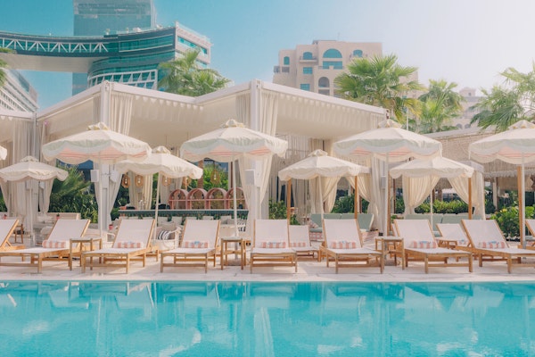 A tranquil pool area with lounge chairs and white cabanas, surrounded by lush greenery and modern buildings, reflecting a relaxing atmosphere.