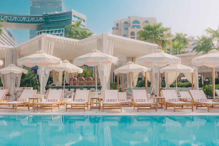 A tranquil pool area with lounge chairs and white cabanas, surrounded by lush greenery and modern buildings, reflecting a relaxing atmosphere.
