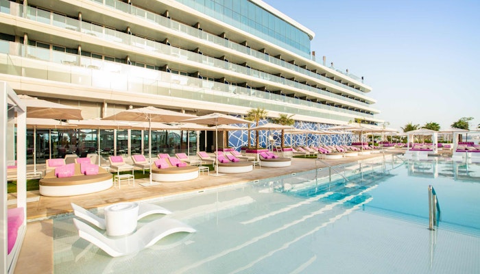 W Dubai The Palm – WET Deck A stylish pool area with modern seating pods and loungers, surrounded by a contemporary building and palm trees.