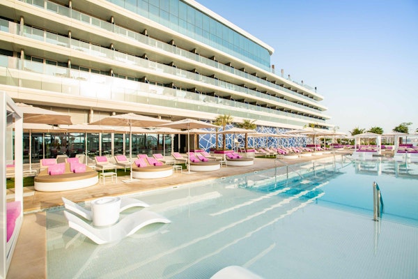A stylish pool area with modern seating pods and loungers, surrounded by a contemporary building and palm trees.