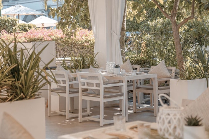 Outdoor dining area with white tables and chairs surrounded by greenery.