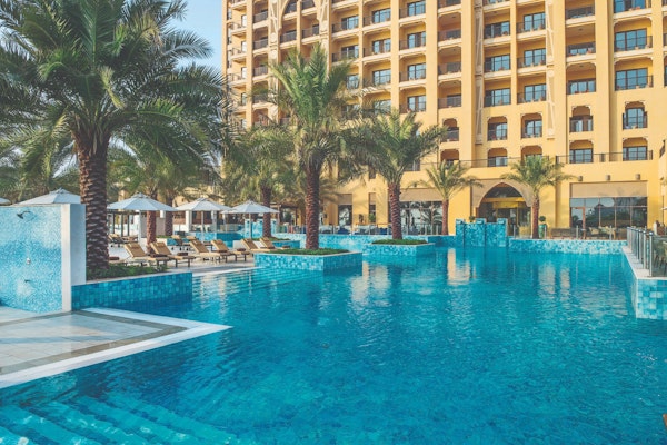 A swimming pool surrounded by palm trees and sun loungers, with a hotel building in the background.