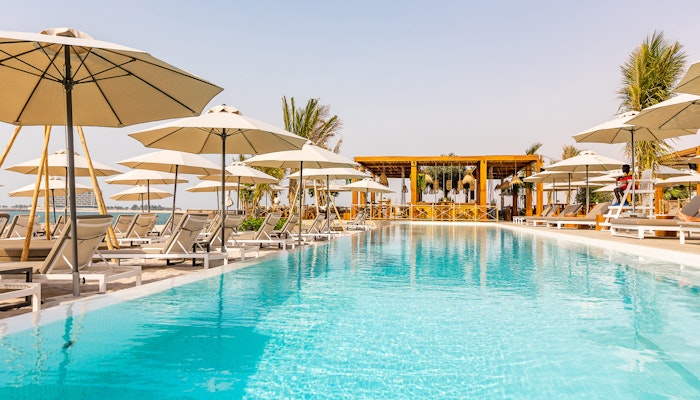 Ula Beach RAK Mövenpick Resort Al Marjan Island A serene poolside view featuring sun loungers and umbrellas, leading to a bar area with a tropical atmosphere.