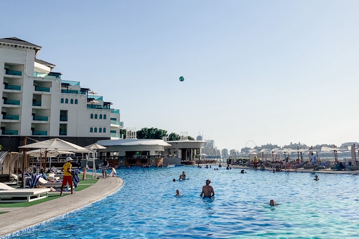 A sunny day at a hotel pool with guests swimming and relaxing; a ball is seen flying through the air