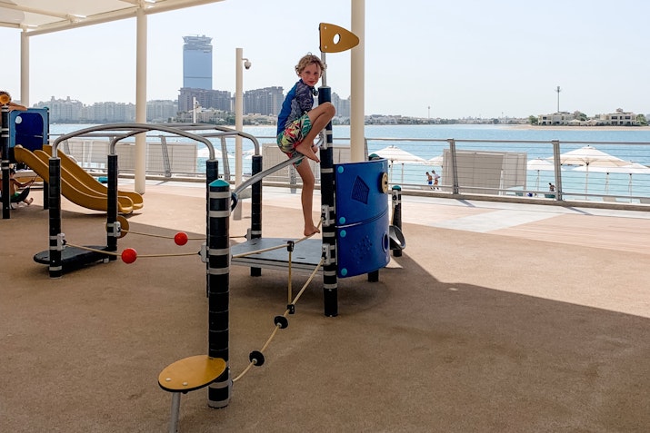 A boy playing on a colorful playground structure with a slide, standing beside it with a waterfront view in the background