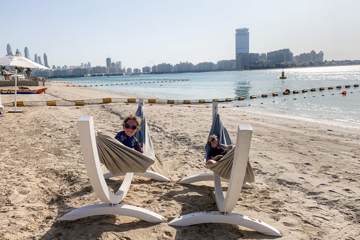 Two children relaxing in beach hammocks, with a view of the waterfront and city skyline in the background