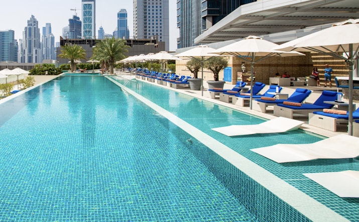 A swimming pool with lounge chairs under umbrellas and city buildings in the background.