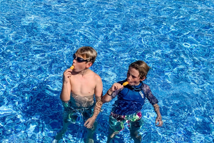 Two boys in sunglasses standing in a swimming pool, each holding a popsicle, with clear blue water surrounding them