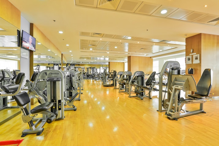Spacious gym interior with various exercise machines on a polished wooden floor, featuring mirrors and television screens