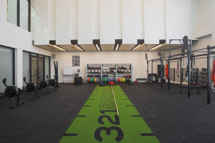 A spacious gym with a green workout track in the center, surrounded by exercise equipment like rowing machines, weights, and functional training gear.