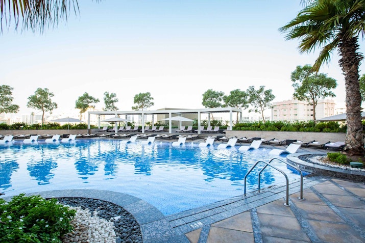 Swimming pool with reclining lounge chairs, surrounded by greenery and palm trees, under a clear sky.