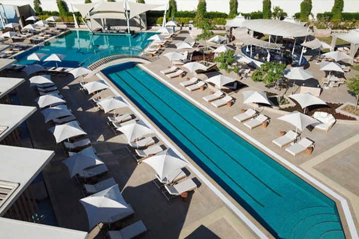 A spacious resort pool with loungers under umbrellas and a separate long lap pool, with a bar area visible in the background.