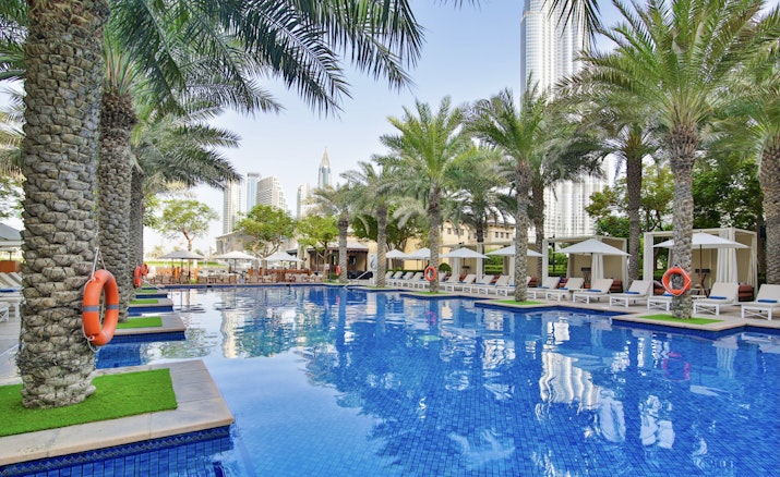 A pool surrounded by palm trees and loungers, with a view of city skyscrapers in the background.