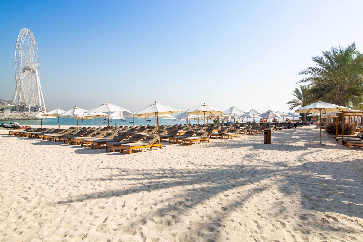 DoubleTree Hilton JBR Rows of wooden beach loungers with umbrellas set on sandy shores, with a view of the ocean and a Ferris wheel in the background.