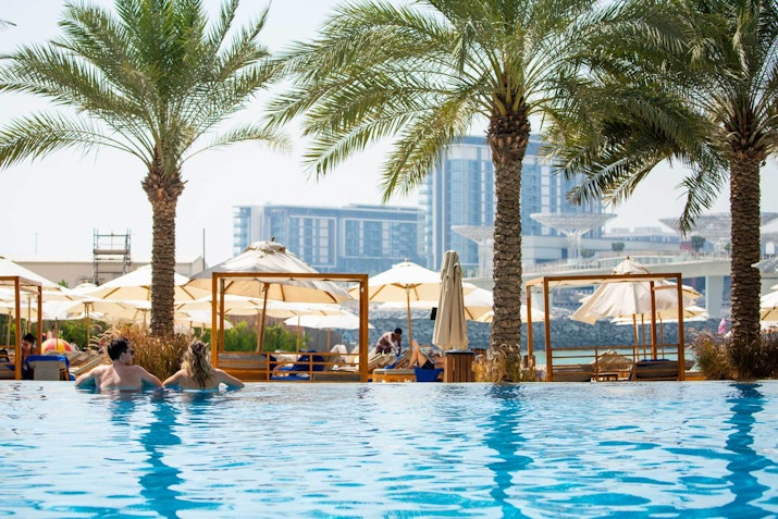 People enjoying a swimming pool surrounded by palm trees and sun loungers, with city buildings in the background