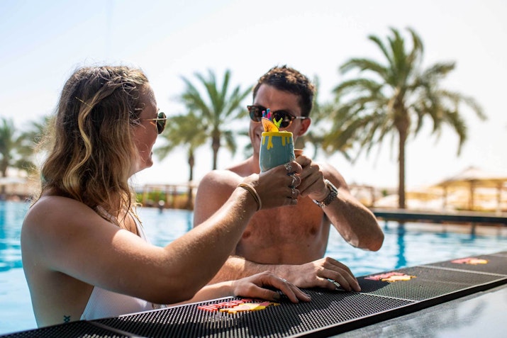 DoubleTree Hilton JBR Couple clinking colorful drinks at a poolside bar, with palm trees and a swimming pool in the background.