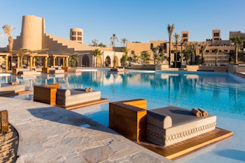 A tranquil pool area featuring lounge beds, surrounded by palm trees and a desert-inspired architectural backdrop.