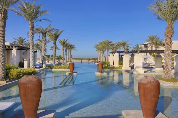 A serene pool area lined with palm trees and large decorative pots, leading to a view of the sea in the distance.
