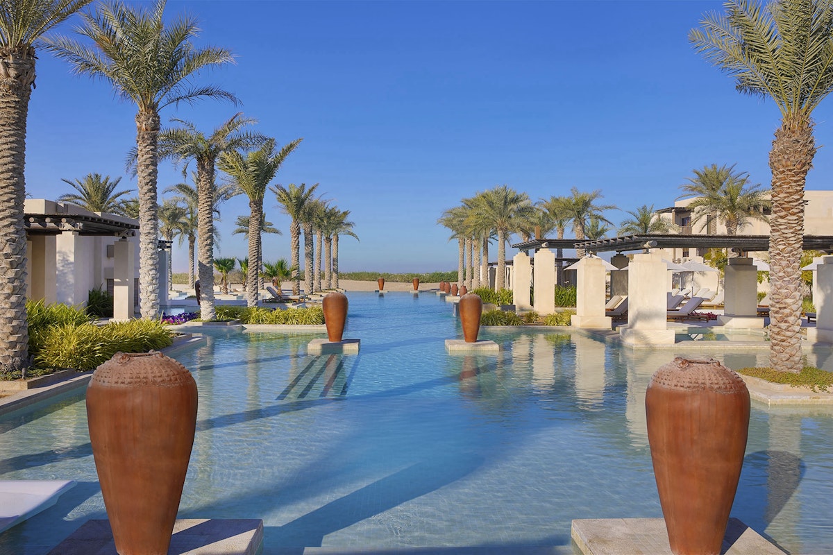 A serene pool area lined with palm trees and large decorative pots, leading to a view of the sea in the distance.