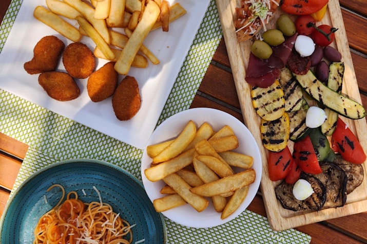 A table spread featuring a variety of dishes, including French fries, croquettes, grilled vegetables, and a pasta dish.