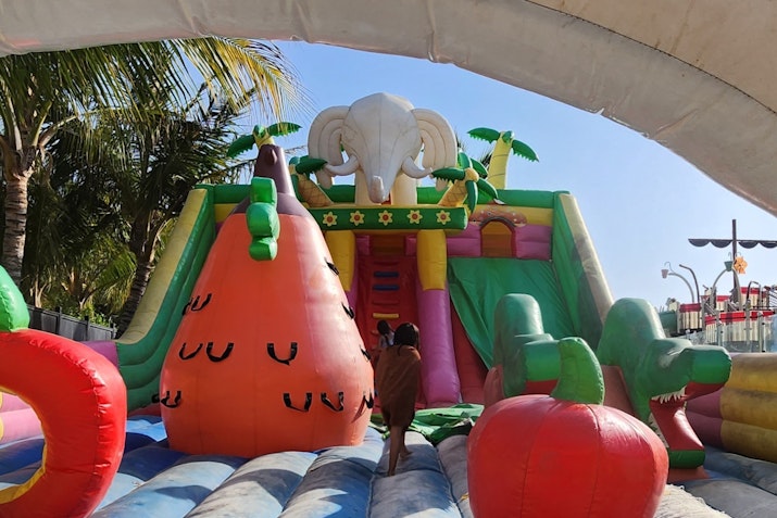 A colorful inflatable playground featuring a slide, an elephant design, and various fruit shapes, with a child climbing up.