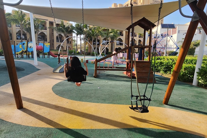 A child swinging on a wooden swing in a playground surrounded by greenery and colorful play structures.
