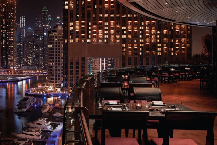Elegant outdoor dining overlooking a marina at night with city lights and boats in view.