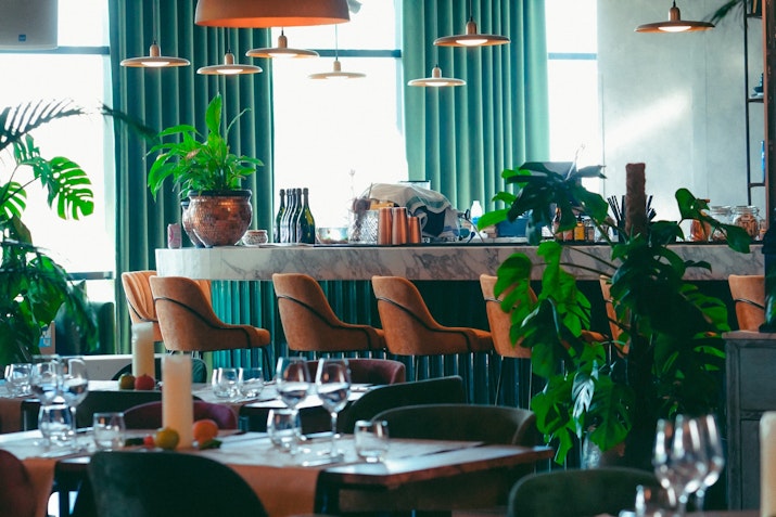 Modern restaurant interior with green curtains, potted plants, and a marble counter with bar seating.