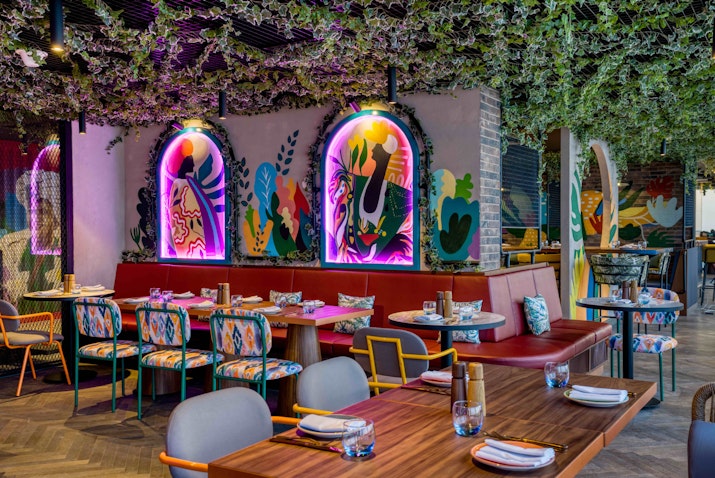Vibrant restaurant interior with colorful wall art, neon lighting, patterned chairs, and greenery-covered ceiling.