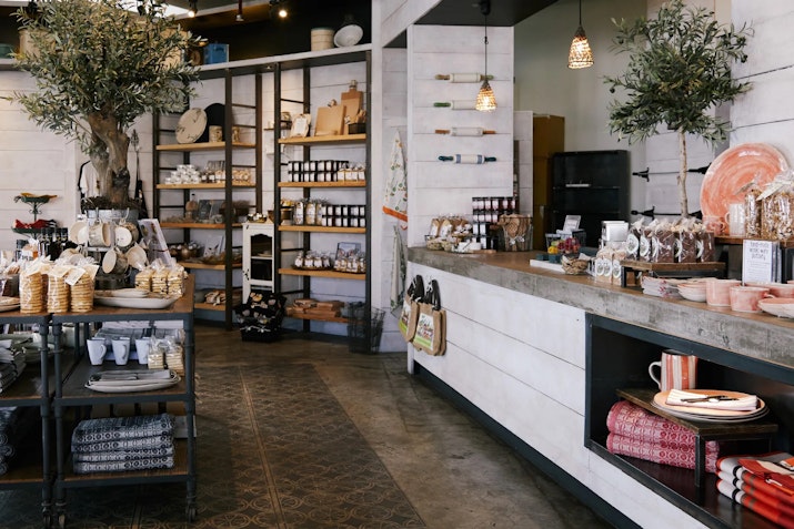 Rustic boutique shop interior with shelves of artisanal goods, ceramics, and decorative items, accented by a potted olive tree