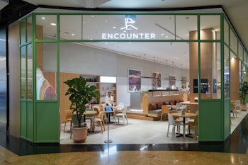 Modern café entrance with green-framed glass walls, minimalist decor, and 'Encounter' sign above, showing seating area inside.