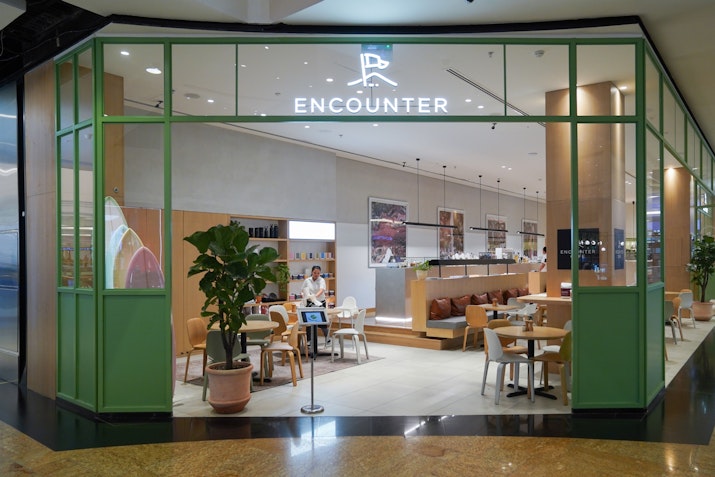 Modern café entrance with green-framed glass walls, minimalist decor, and 'Encounter' sign above, showing seating area inside.