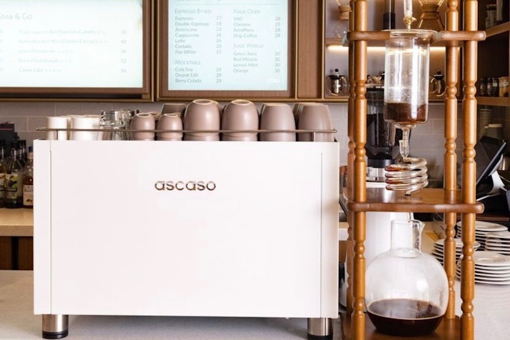 White Ascaso espresso machine with stacked cups beside a glass cold brew coffee drip tower on a counter