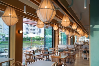 A restaurant featuring large floor to ceiling windows, natural light and wooden seating with unique lighting overhead overlooking lush greenery