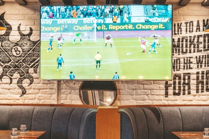 TV displaying a soccer match in a restaurant with mural-decorated brick walls and dark seating booths.