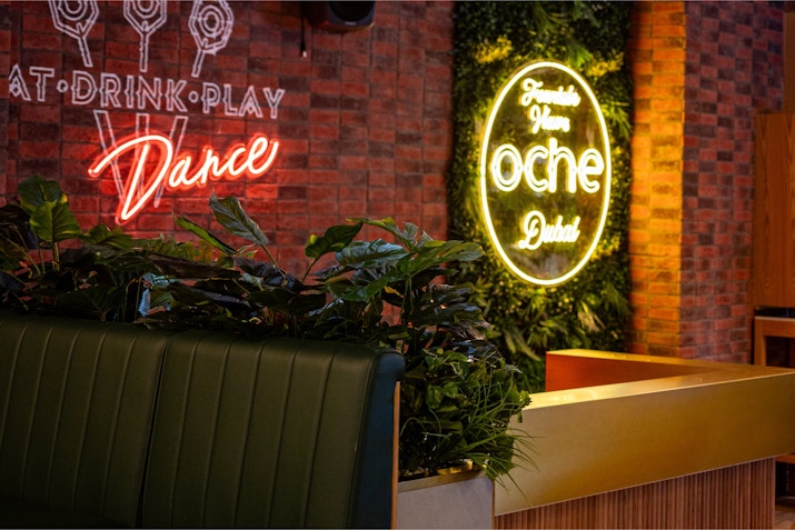 Neon-lit bar area with signs reading 'Eat, Drink, Play, Dance' and 'Oche Dubai,' surrounded by lush greenery.