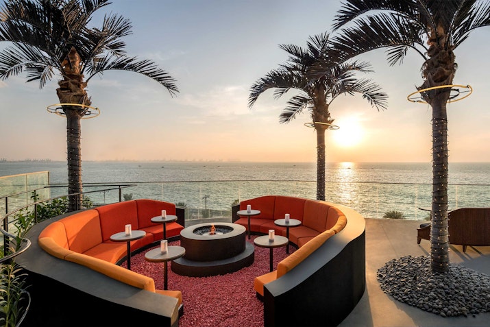 Outdoor seating area with circular couches, a fire pit, and palm trees, overlooking a sunset by the ocean.