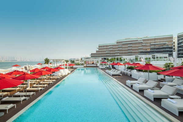 TH8 Palm Dubai Beach Resort - Vignette Collection Large pool with red umbrellas and sun loungers, overlooking the sea.
